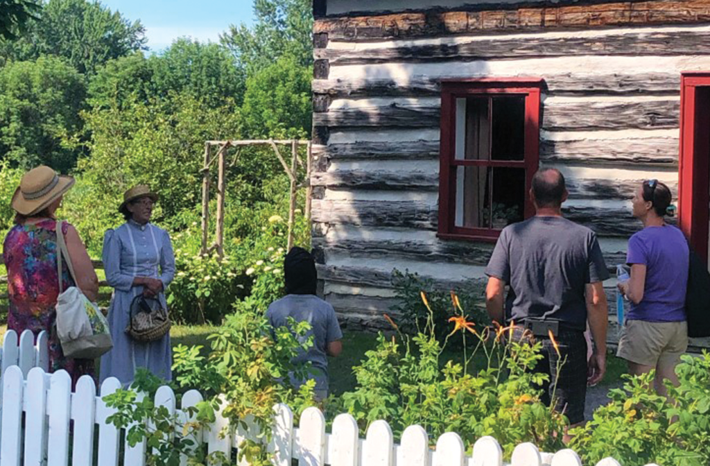Family standing outside of a log cabin listening to tour guide dressed in period clothing